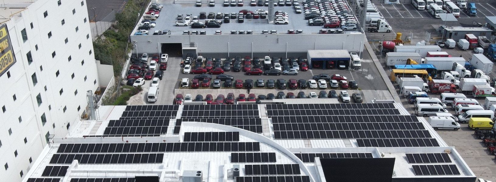 Techo de una agencia automotriz cubierto de filas de paneles solares, con el estacionamiento de autos detrás, vista desde dron