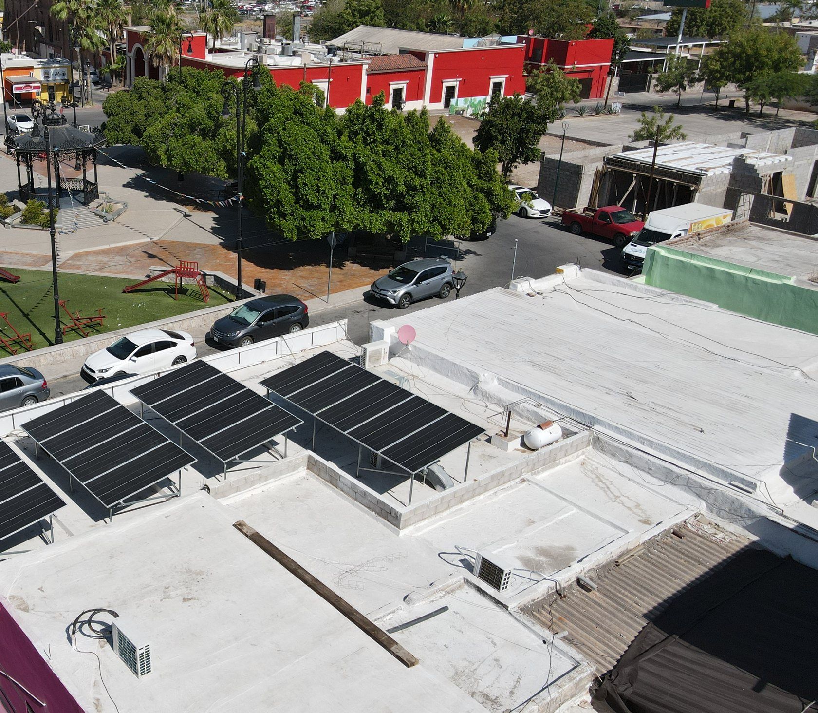 Comercio de barrio en Hermosillo con paneles solares sobre estructuras inclinadas en la azotea, frente a una plaza con árboles, visto desde dron