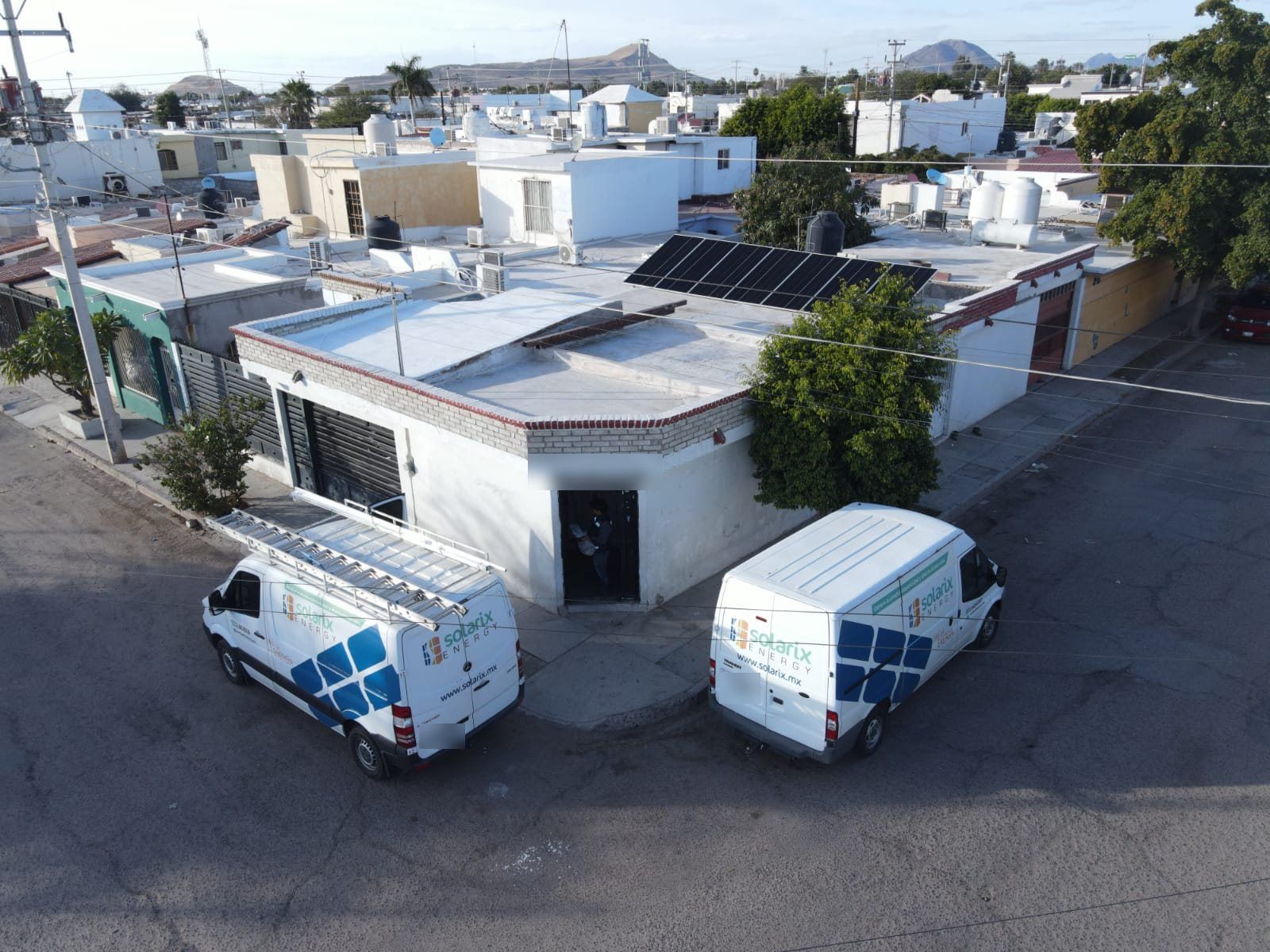 Dos camionetas blancas de Solarix Energy estacionadas frente a un comercio de esquina en Hermosillo con paneles solares en la azotea, vistas desde dron